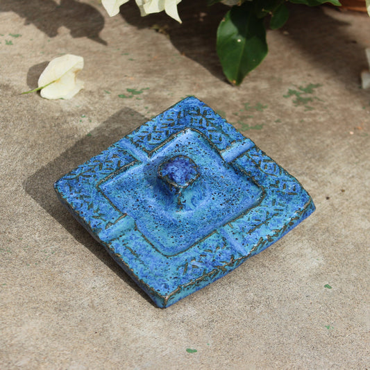 A square blue ceramic dish with a raised center and decorative border, placed on a textured concrete surface with white flowers and green leaves in the background.