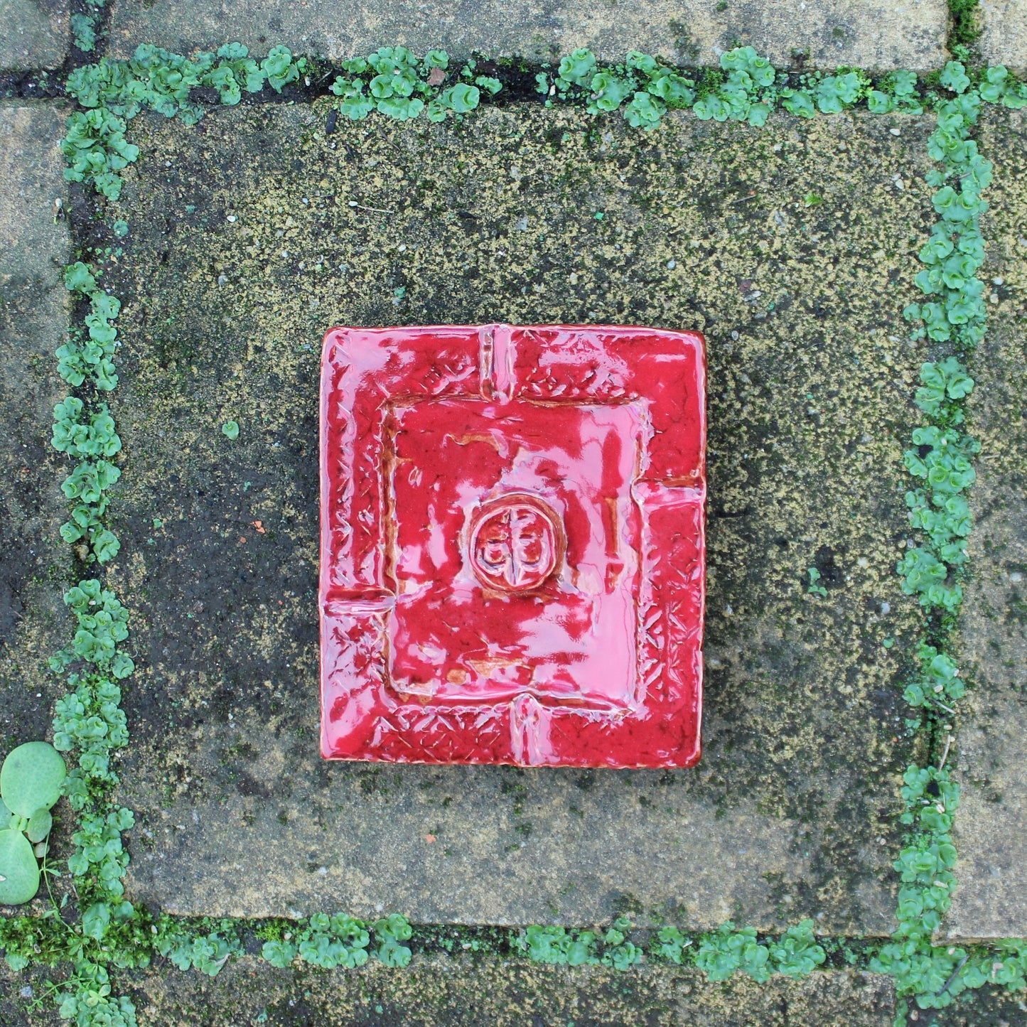 A square red ceramic tile with a raised circular emblem in the center, placed on a weathered stone paver surface with green moss and small plants growing between the stones.